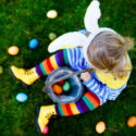 Close-up of legs of toddler girl with colorful stockings and shoes and basket with colored eggs. Child having fun with traditional Easter eggs hunt, outdoors. Unrecognizable face, no face